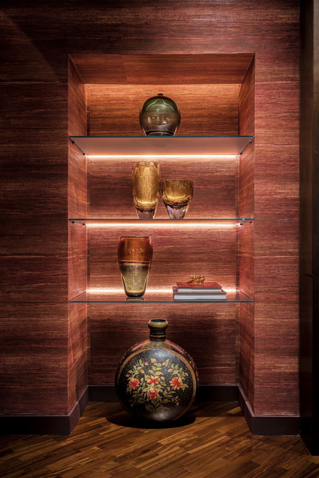 Lit niche in a wood-panelled wall, with glass vases and a painted vase on glass shelves