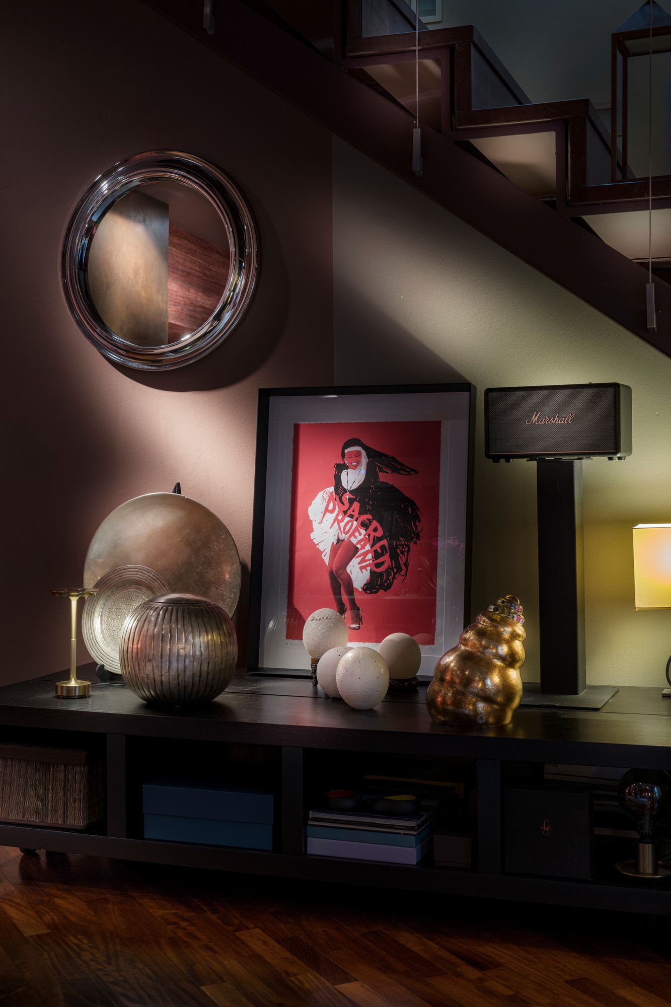 Collected objects on a low cabinet beneath the stair: a round mirror, a framed print and brass pieces